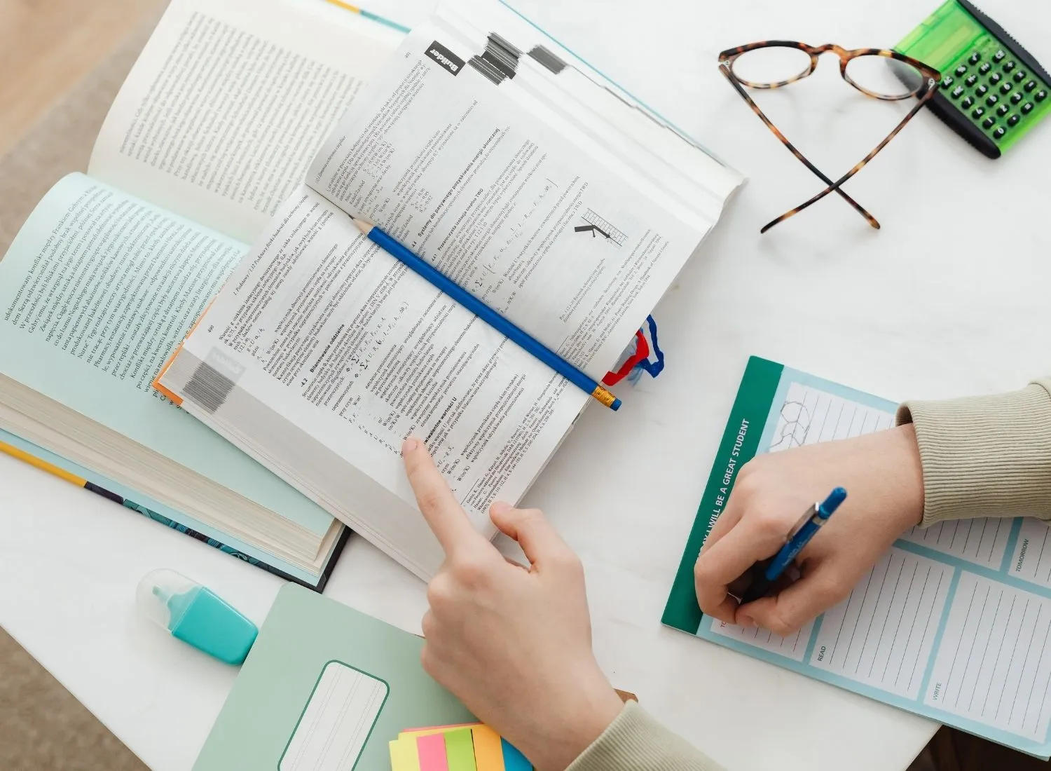 Person studying with open books, pointing at text, writing on planner, with glasses and calculator nearby.