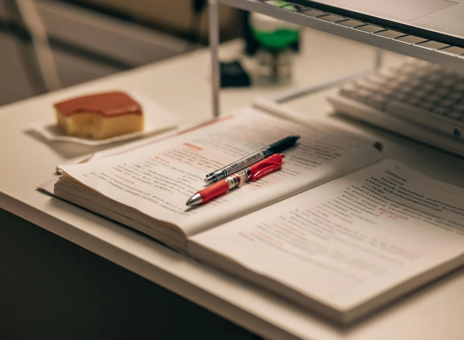Open book with Chinese text and two pens on desk near keyboard and a plate with a pastry.