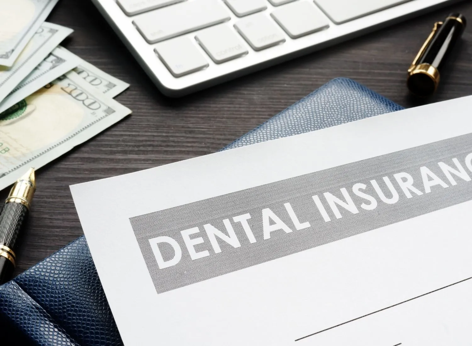 Close-up of dental insurance paper, pen, cash, keyboard, and wallet on dark wooden desk.
