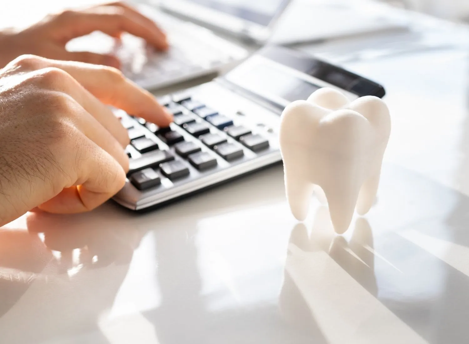 Hands typing on a calculator with a large white tooth model on a reflective surface nearby.