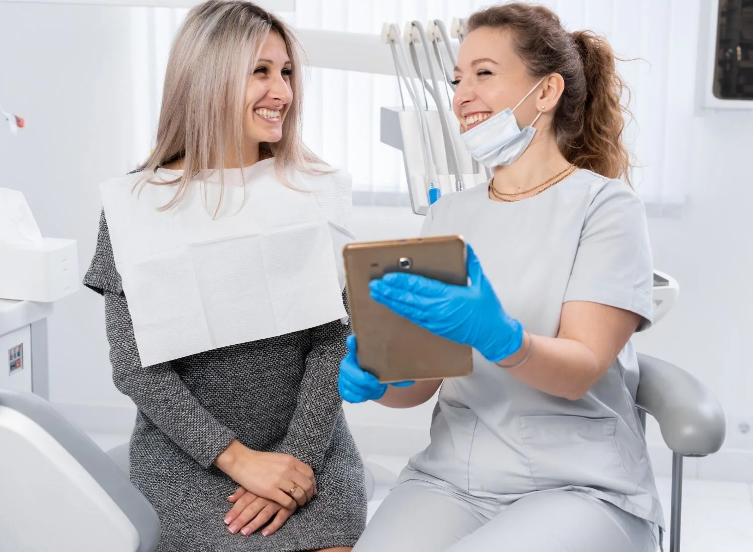 Dentist wearing gloves showing a tablet to a smiling female patient in a dental office.