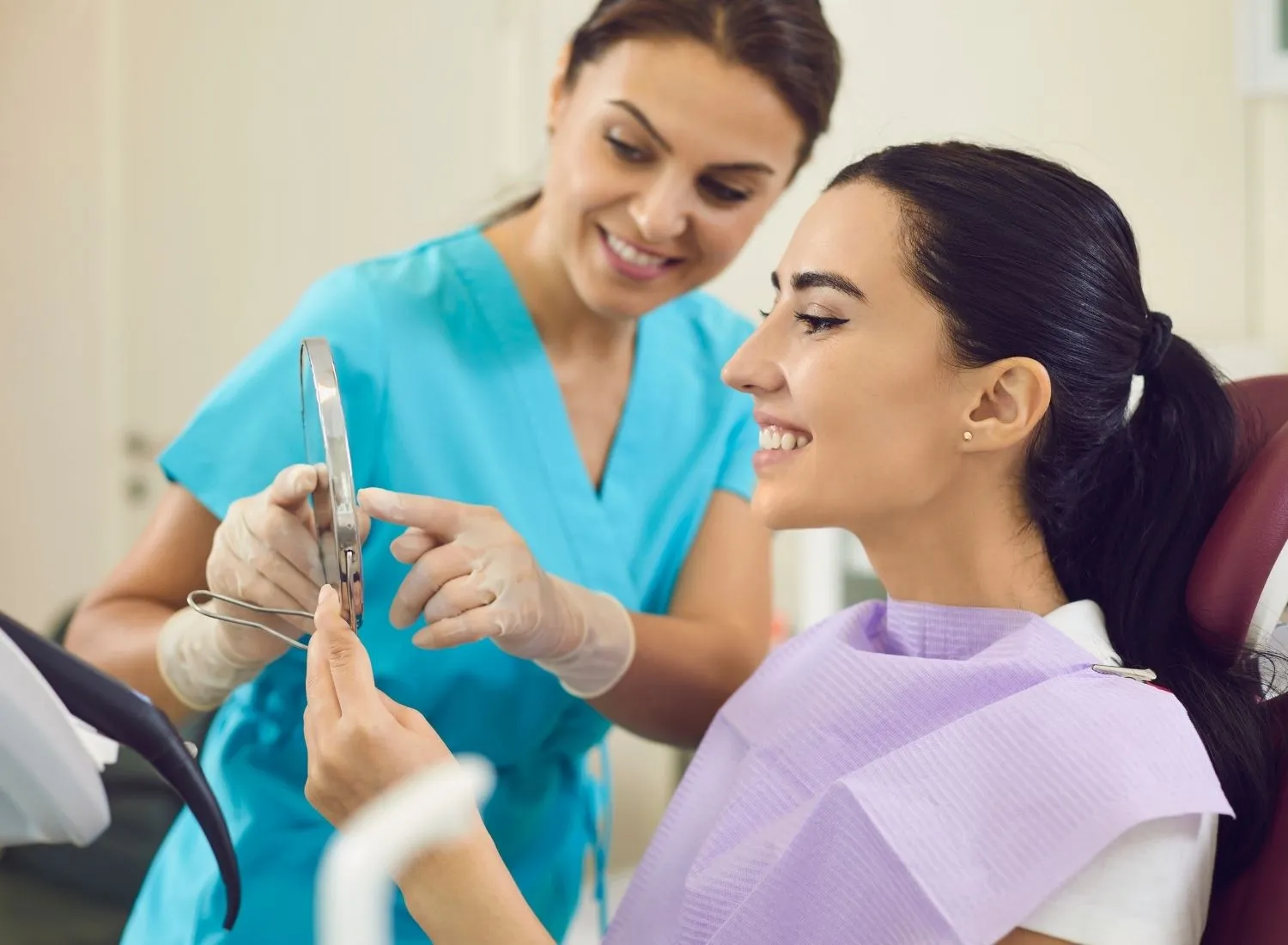 Smiling female dentist in blue scrubs shows a dental mirror to a happy woman in a dental chair.