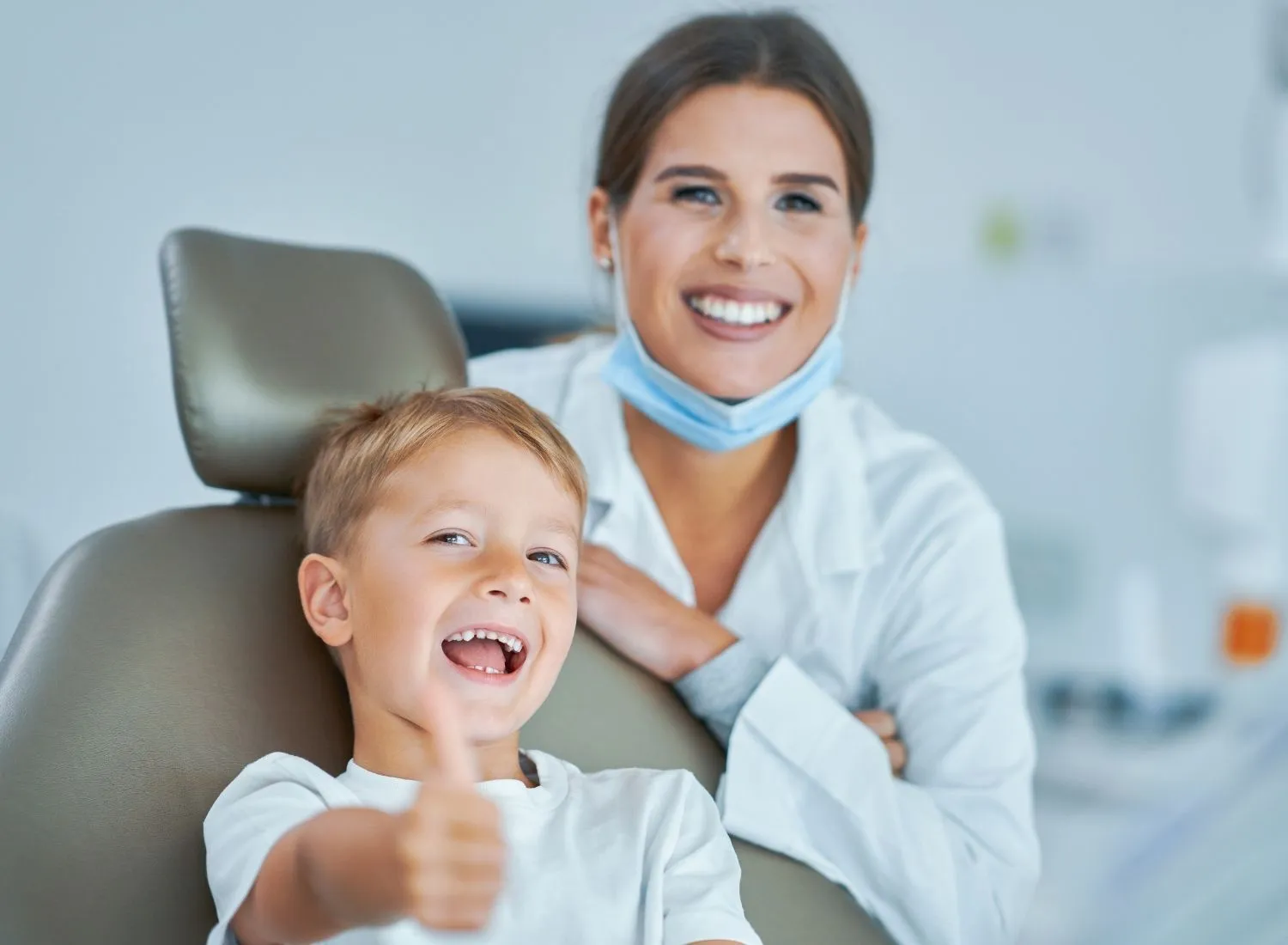 Smiling young boy in dental chair showing thumbs up next to female dentist in mask and white coat.