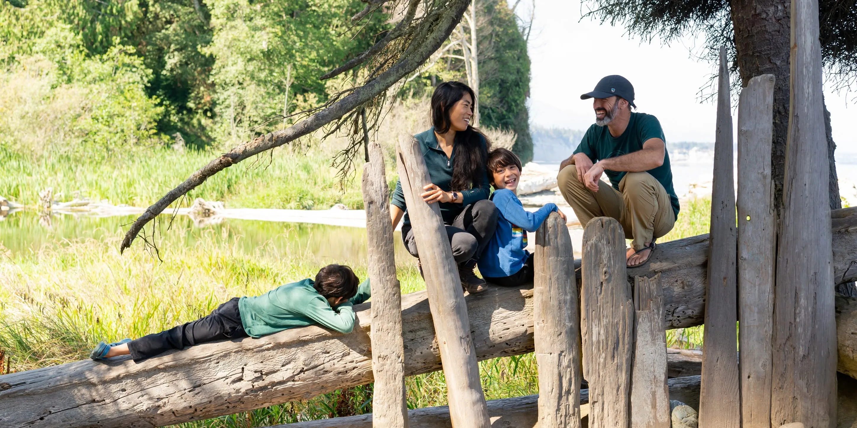 A family of four outdoors sitting and lying on a large fallen tree trunk with greenery and a body of water in the background, sharing a relaxed and happy moment together.