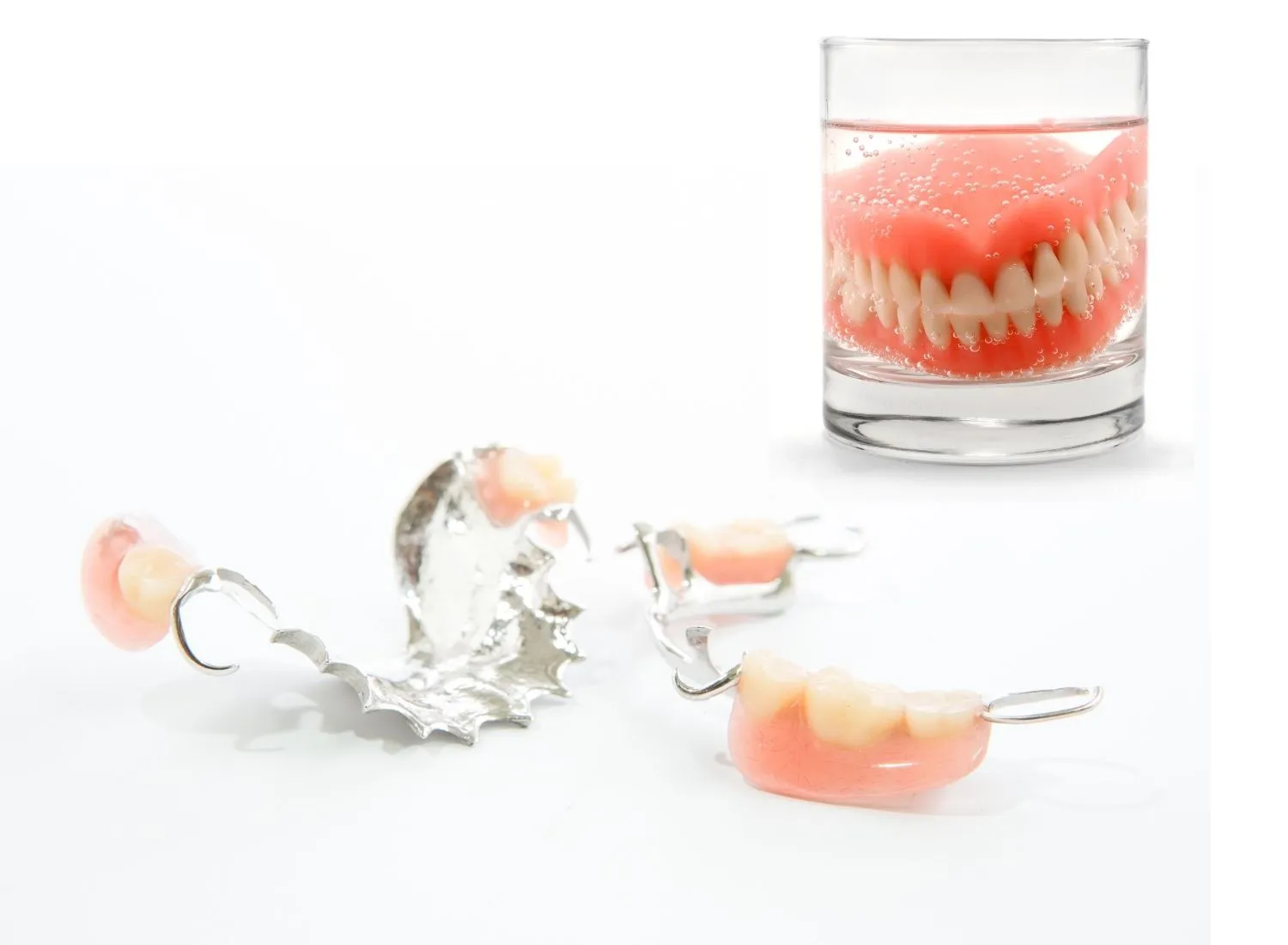 Fragmented partial dentures with metal clasps and artificial teeth arranged on a white surface, alongside a full set of dentures soaking in a clear glass of water with bubbles.