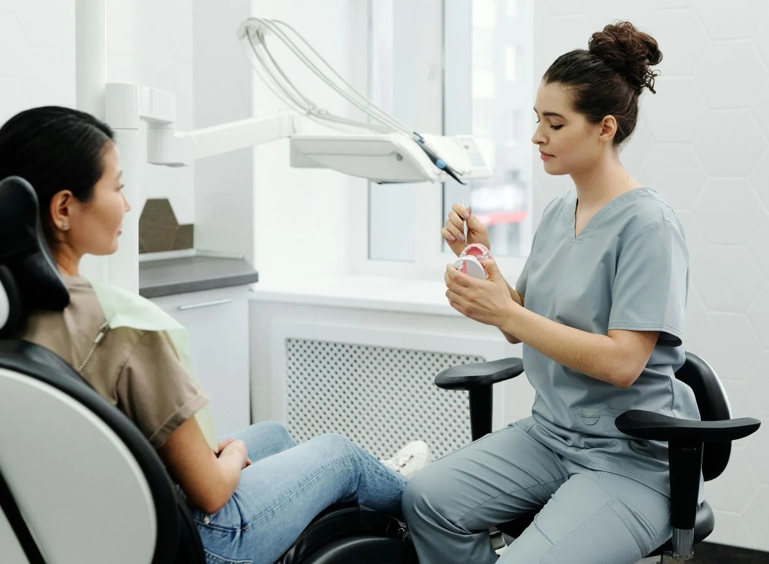 Dentist in scrubs explaining dental care using a mouth model to a seated patient in a clinic room with dental equipment.