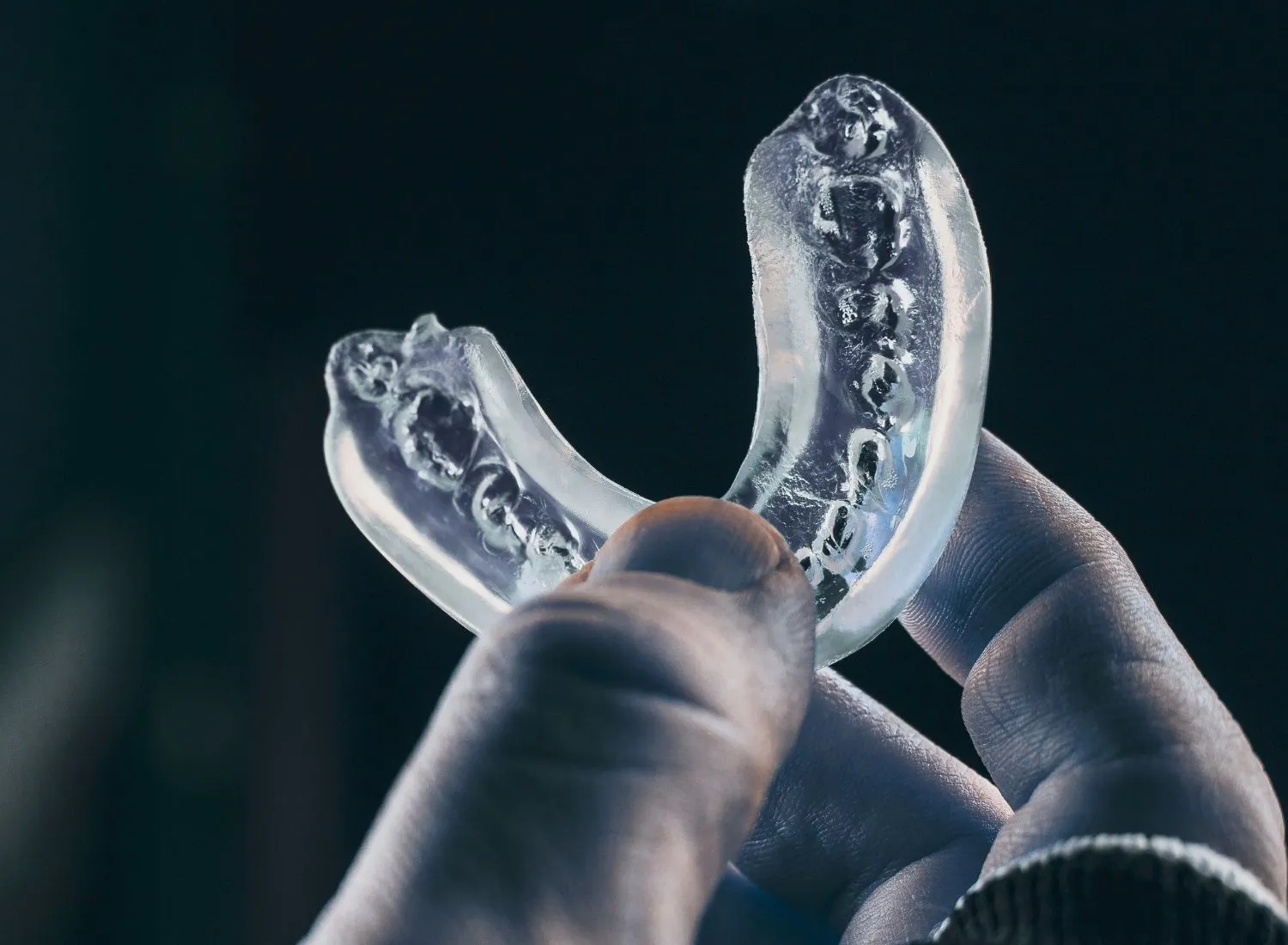 Close-up of a hand holding a transparent dental night guard used for teeth grinding protection, shown against a dark background.