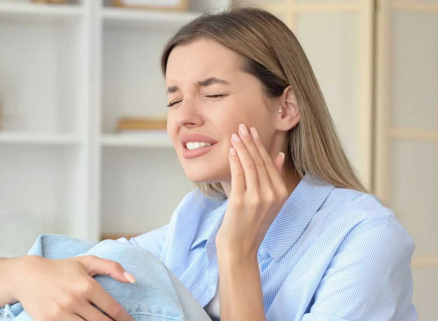 Young woman with long light brown hair in a striped blue shirt sitting and holding her jaw in pain with eyes closed, indicating toothache.
