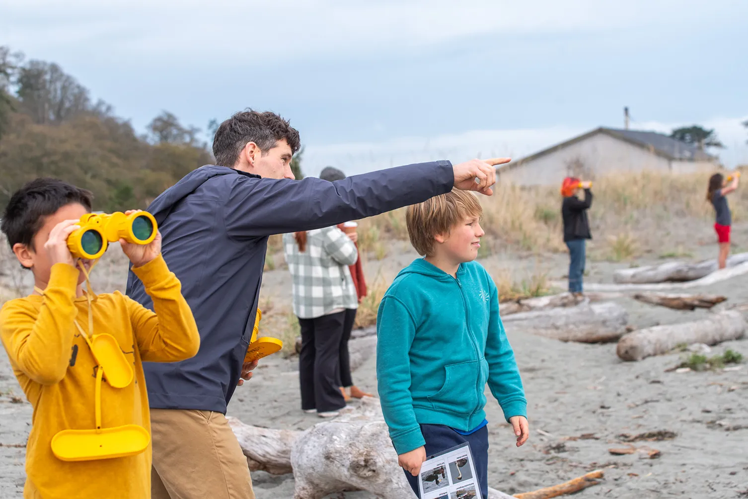 A man points into the distance on a beach while a boy in a turquoise hoodie holds a sheet and looks ahead, and another boy in a mustard yellow shirt looks through yellow binoculars; in the background, two other children use binoculars near tall grass with logs scattered on the sand.