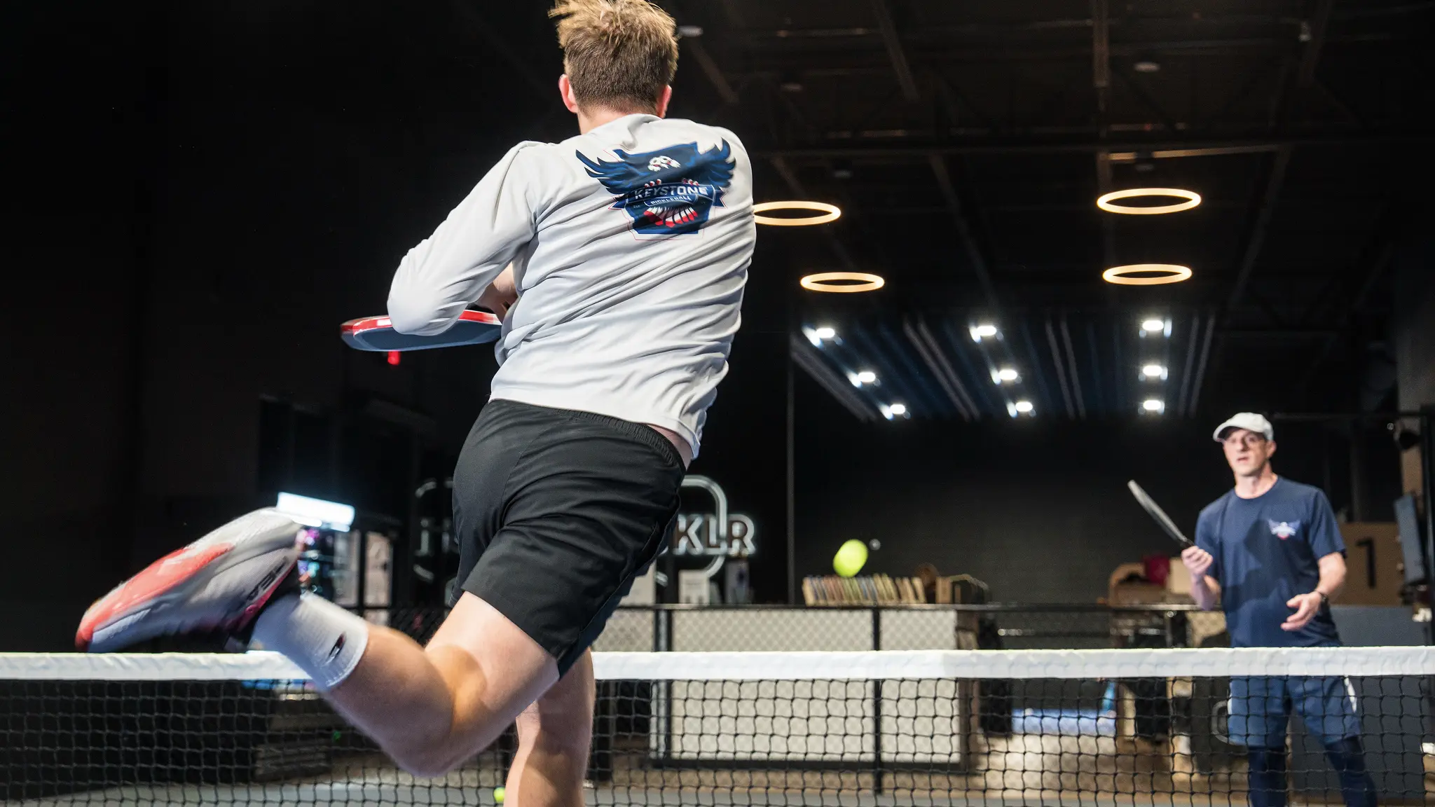 Two men playing pickleball indoors with one mid-jump hitting the ball across the net, wearing a Keystone Pickleball shirt