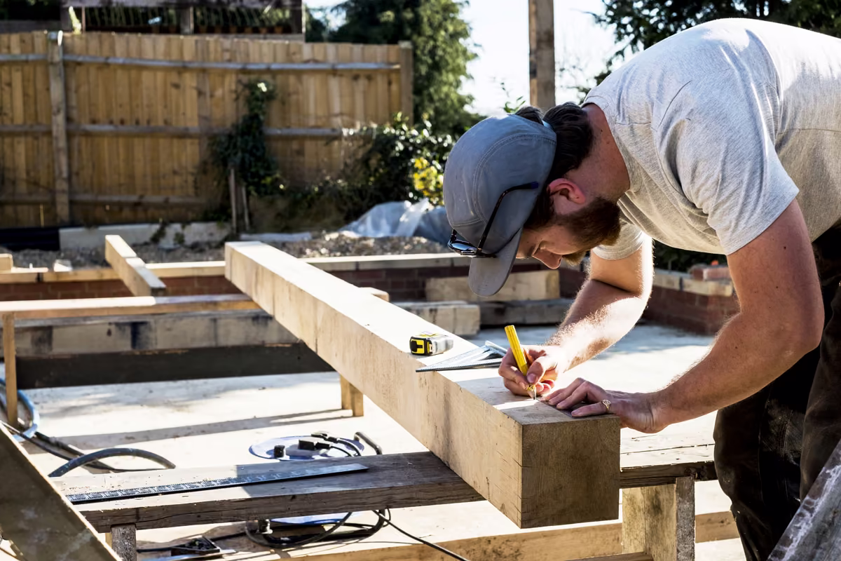 Man in gray shirt and cap marking measurements on a large wooden beam at a construction site.