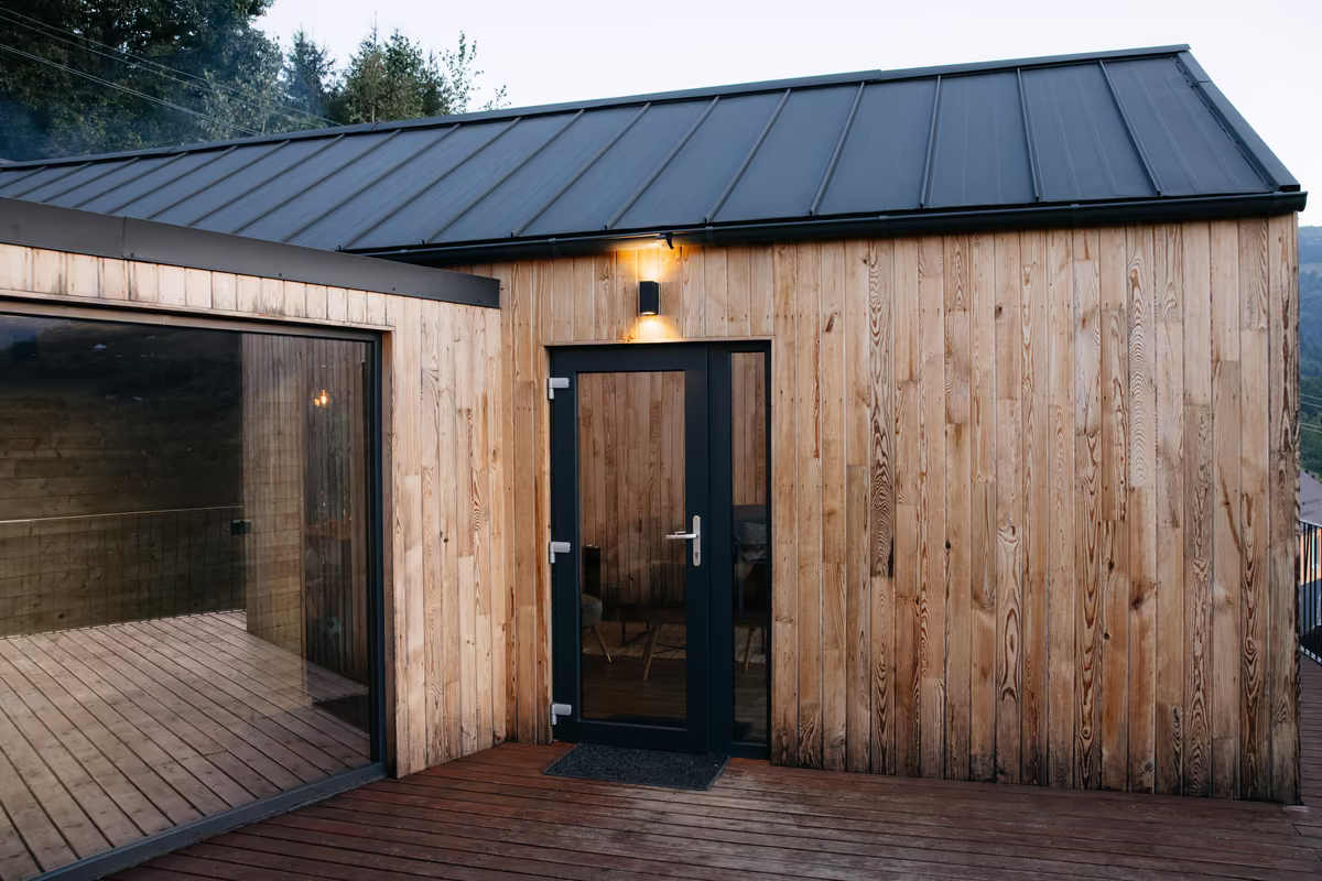 Wooden cabin exterior with glass door, large window, and black metal roof in a wooded area.
