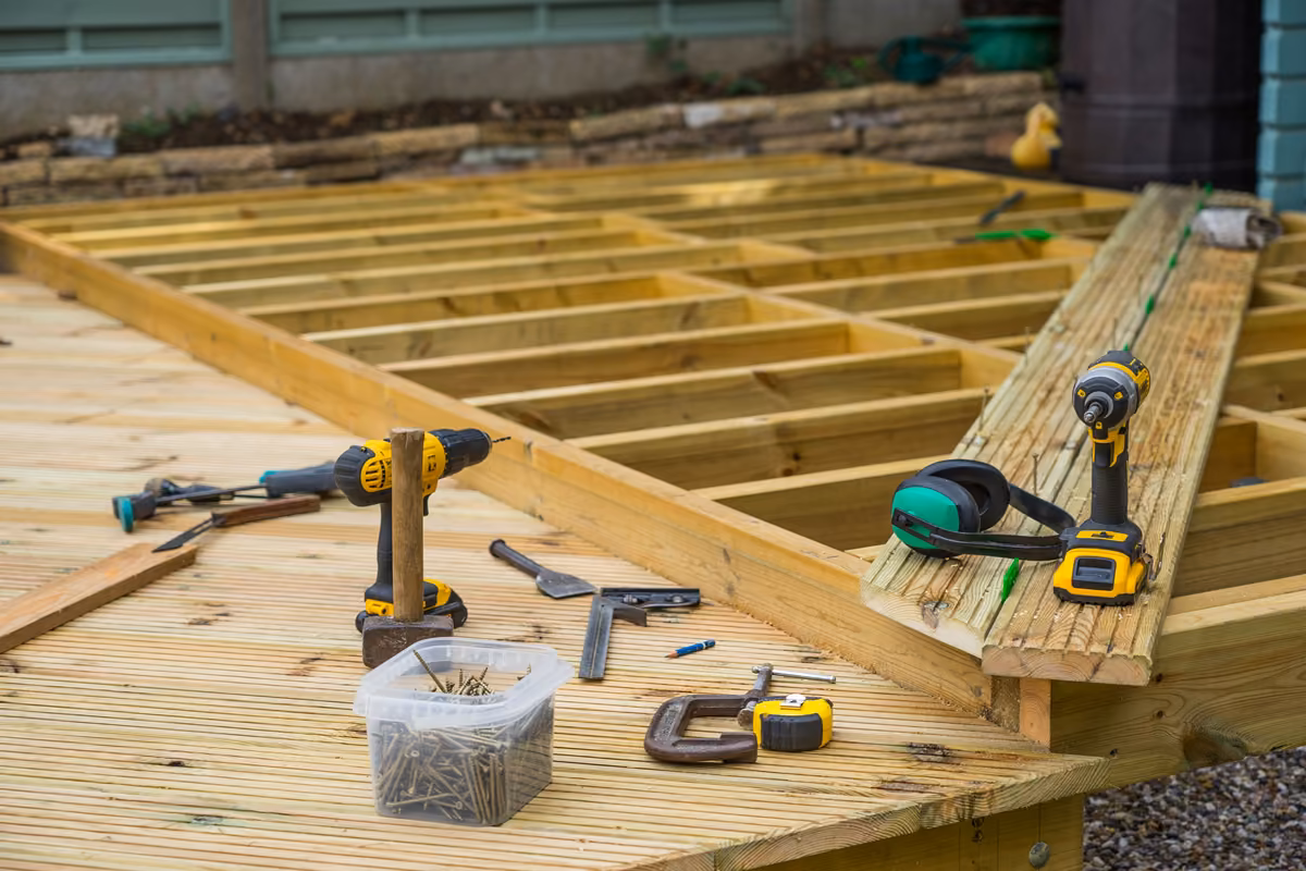 Partially built wooden deck with power drills, screws, clamp, and ear protection on the boards.
