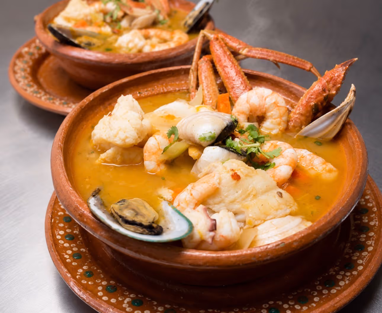 Two clay bowls of seafood soup with shrimp, crab legs, clams, fish, and herbs in broth.