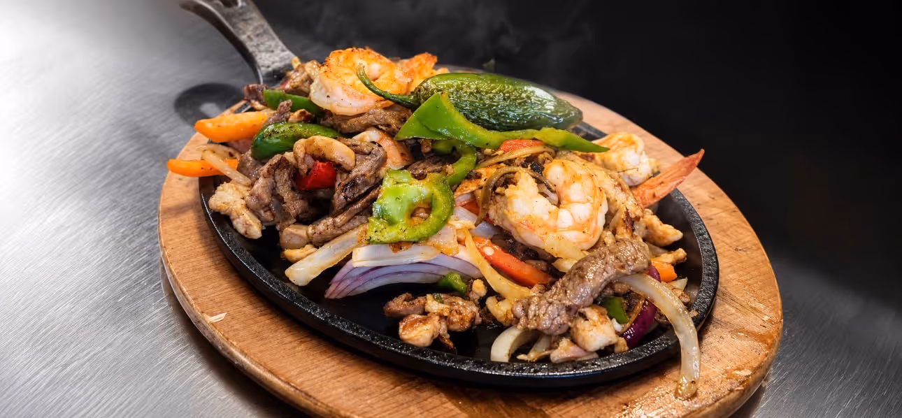 Sizzling skillet of mixed fajitas with shrimp, beef, bell peppers, onions, and jalapeño on wooden board.