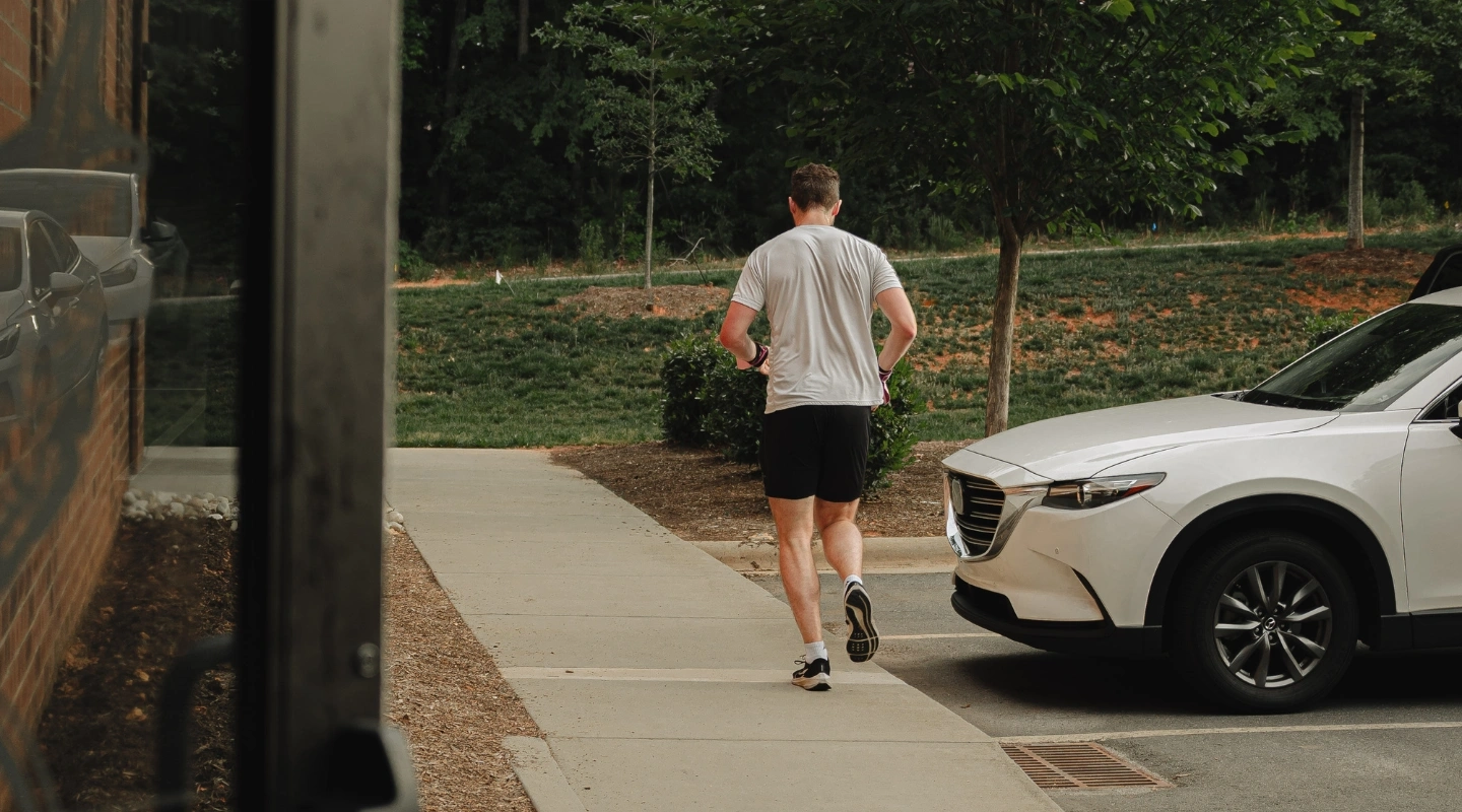 A man in athletic wear jogs away from the camera down an outdoor sidewalk next to a white SUV.