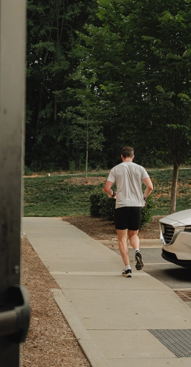 A man in athletic wear jogs away from the camera down an outdoor sidewalk next to a white SUV.