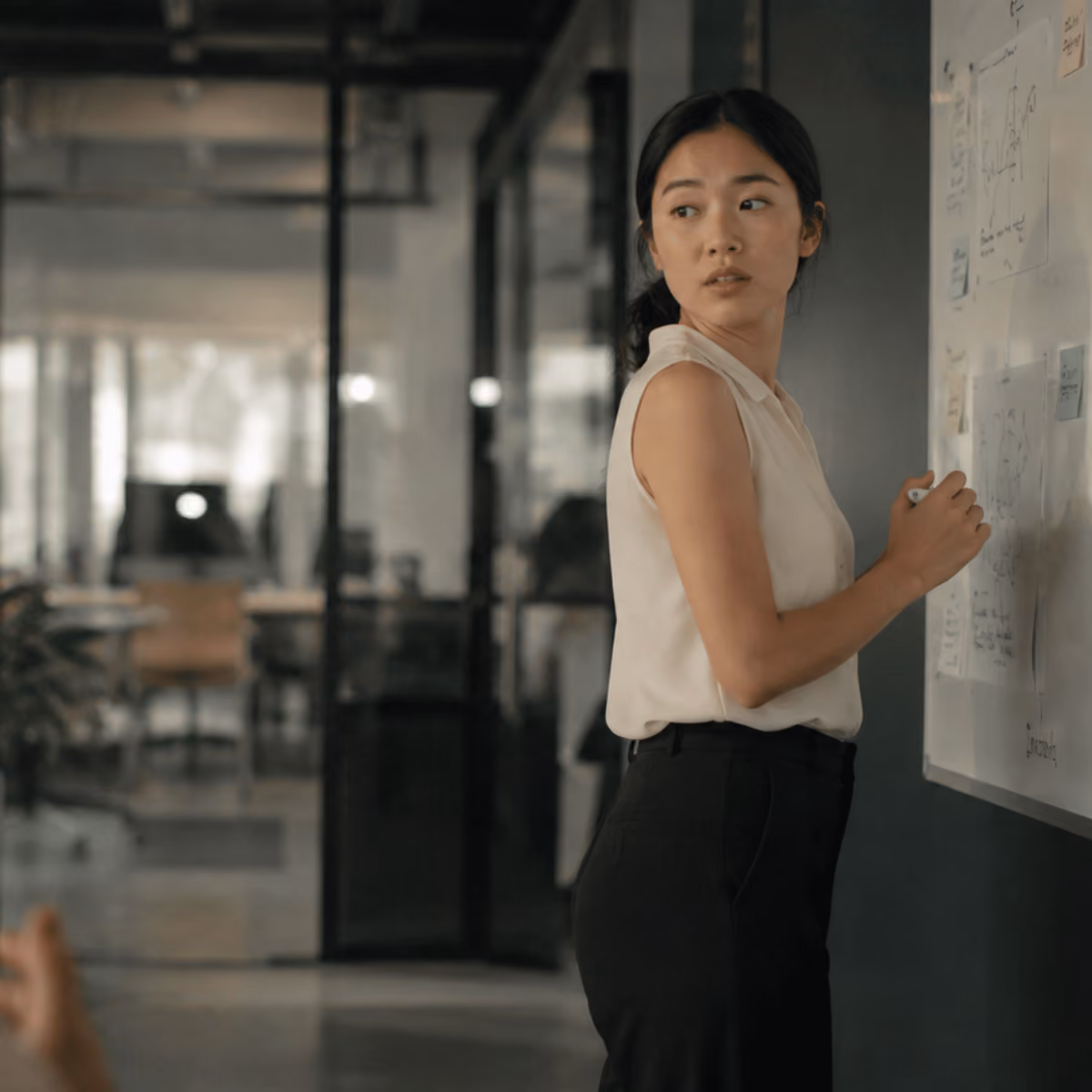 Young woman in business casual writing on a whiteboard in a modern office.