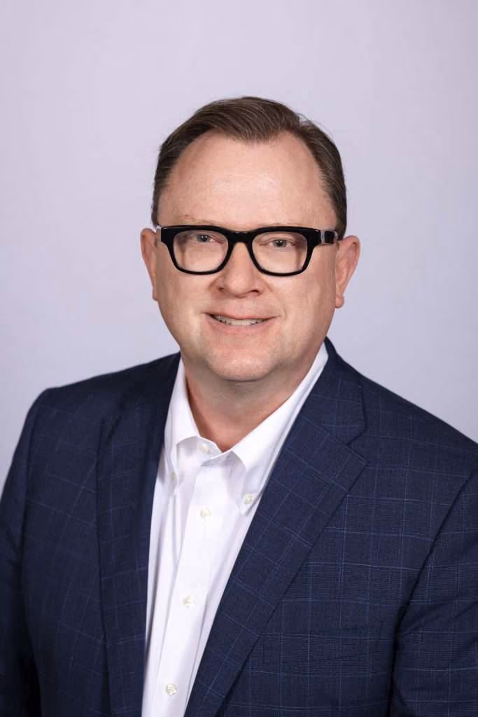 Man with dark hair and glasses wearing a white shirt and blue plaid blazer, smiling at the camera.