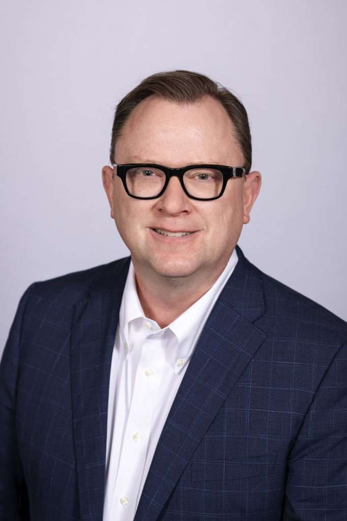 Man with dark hair and glasses wearing a white shirt and blue plaid blazer, smiling at the camera.