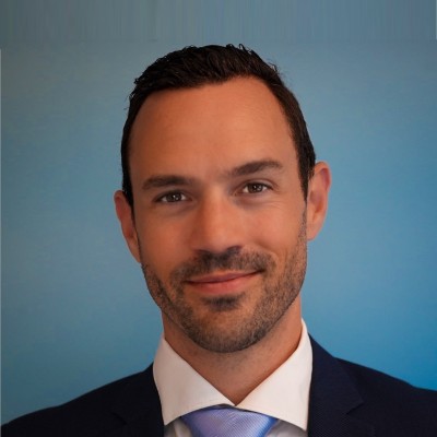 Smiling man with short dark hair and beard wearing suit, white shirt, and light blue tie against blue background.