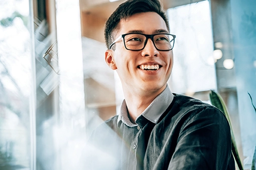 Smiling young man wearing glasses and a dark shirt in a bright indoor setting near a window.