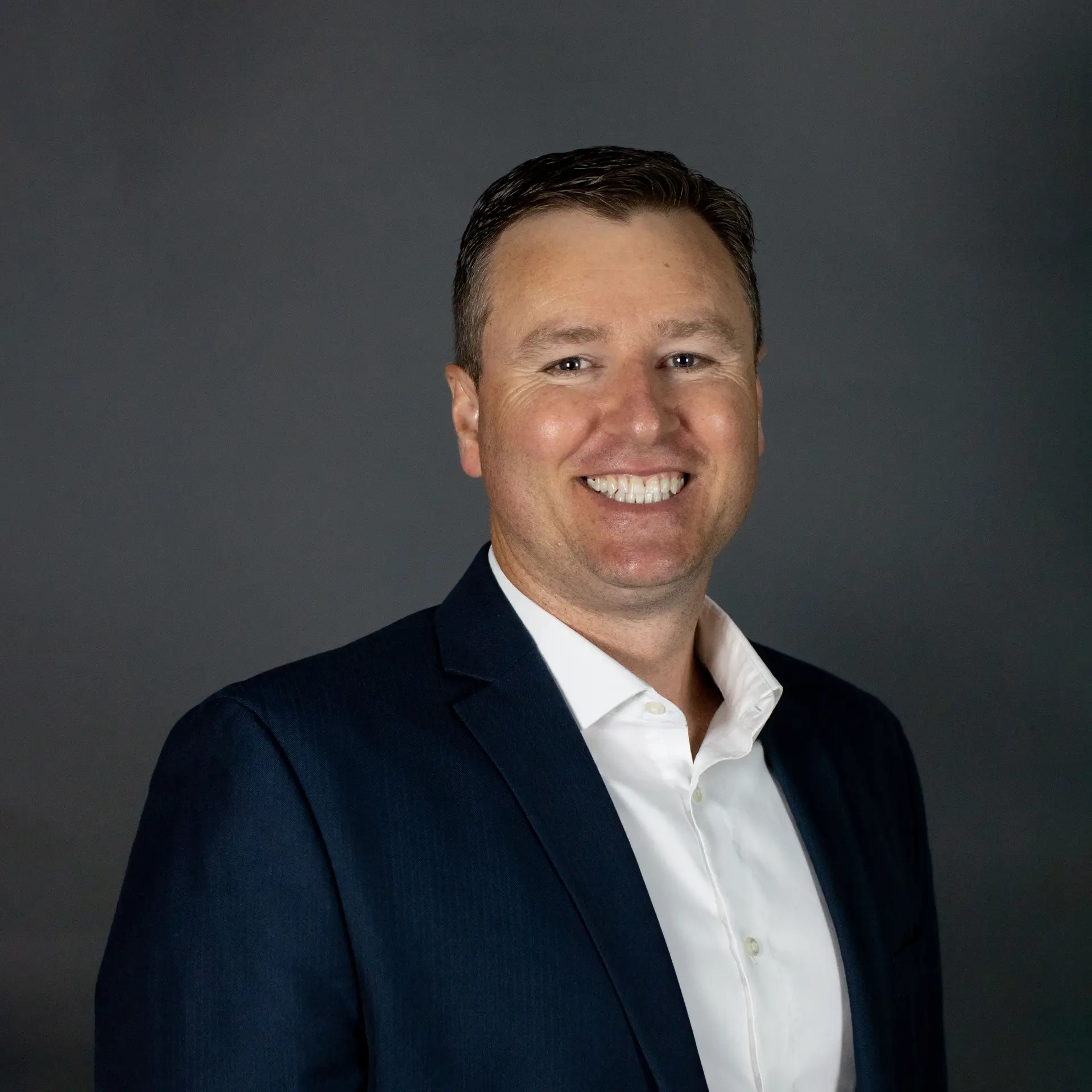Smiling man wearing a dark suit jacket and white shirt against a plain grey background.