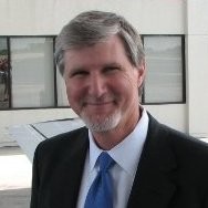 Smiling man with gray hair and beard wearing a suit and blue tie outdoors near building windows.