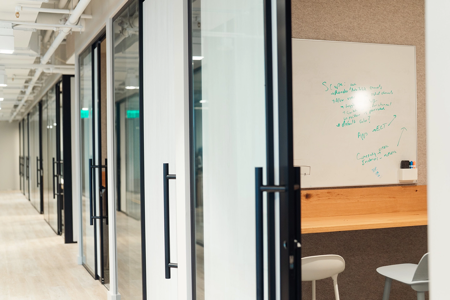Modern office hallway with glass doors and a small meeting room with whiteboard and chairs.