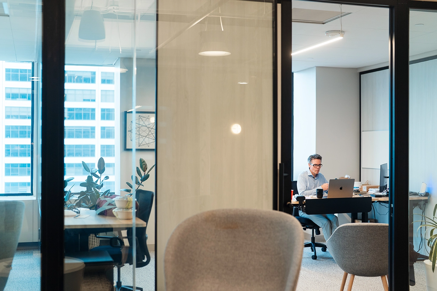 Man working on laptop in a modern office with glass walls and gray chairs.