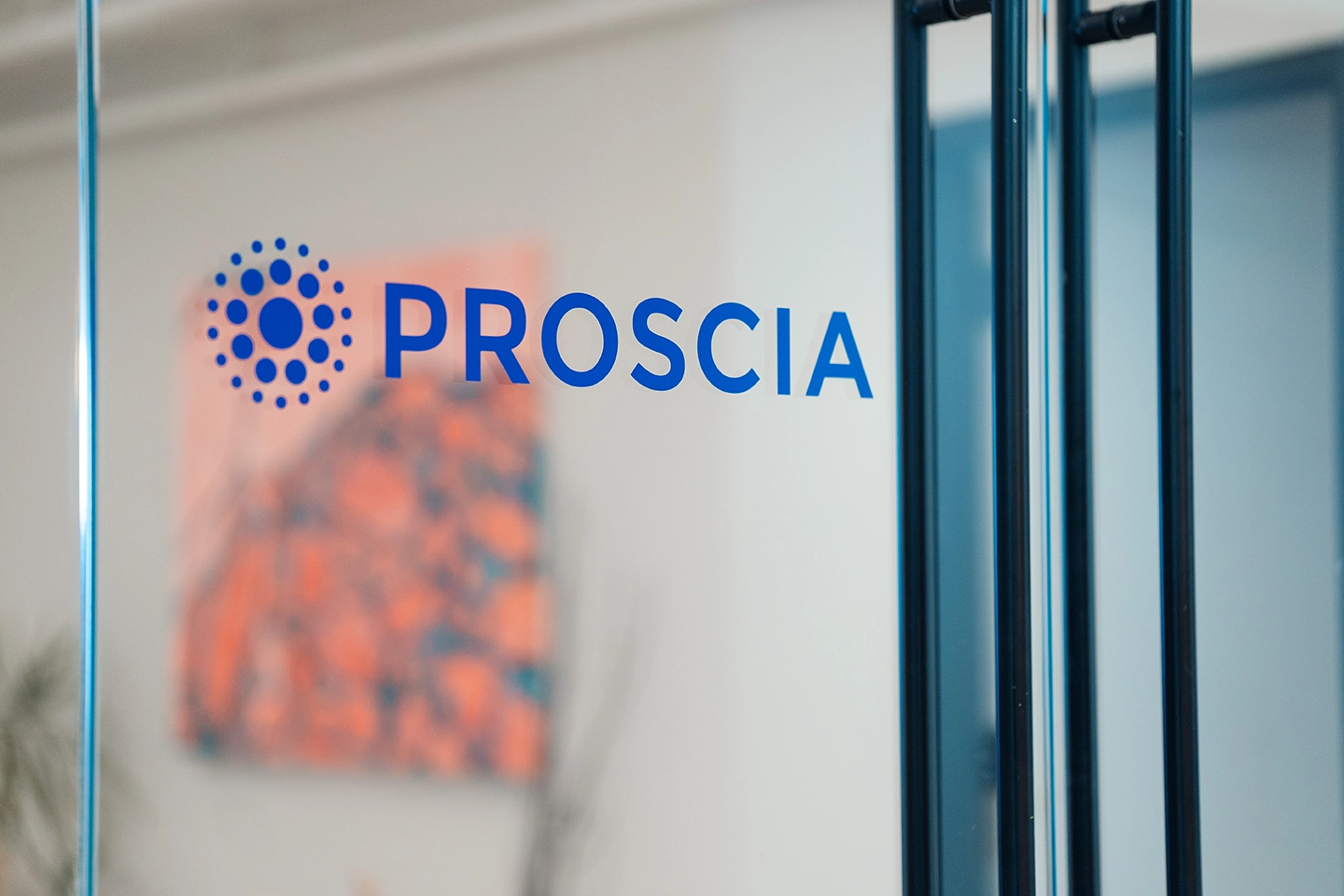 Blue PROSCIA logo with circular dot design on glass office door with blurred background.