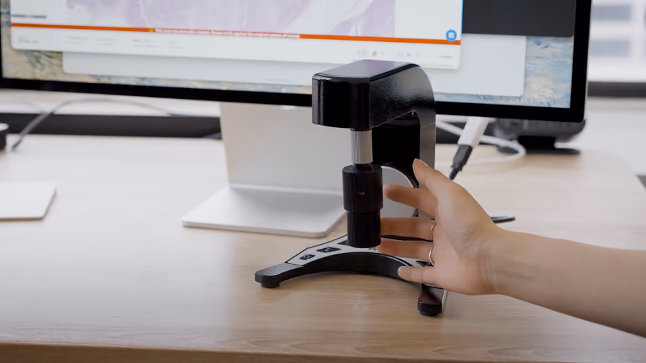A person's hand adjusting a small black measuring device on a wooden desk in front of a computer monitor showing a partly visible map or graph.