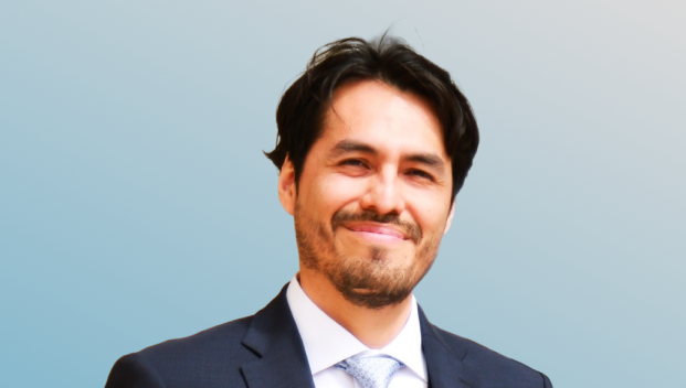 Smiling man with dark hair and beard wearing a dark suit, white shirt, and light blue tie against a blue gradient background.