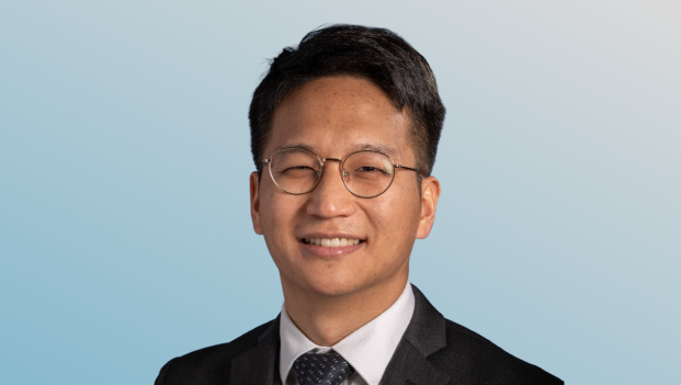 Portrait of a smiling young Asian man wearing round glasses, a black suit, white shirt, and a dark patterned tie, with a gradient light blue to gray background.