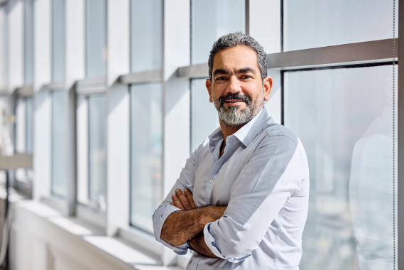 Middle-aged man with gray hair and beard wearing a white dress shirt standing with arms crossed by a large window in a bright office space.