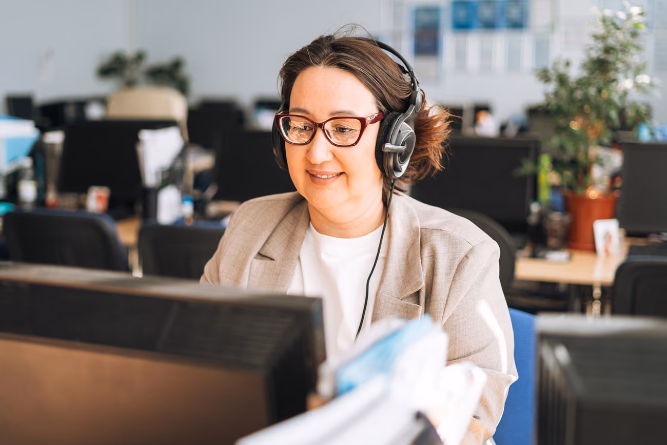 Woman wearing glasses and a headset smiling while working at a computer in an office.