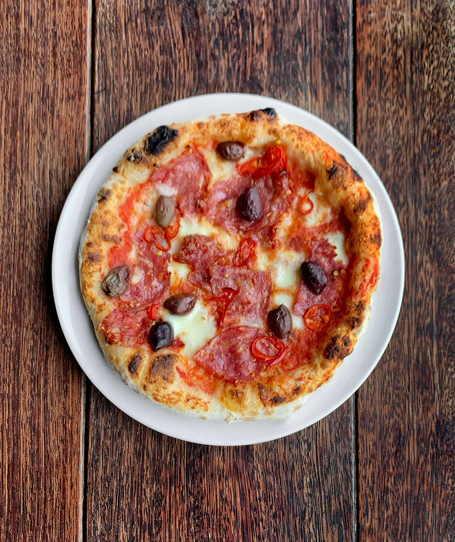 Pizza with tomato, melted cheese, salami slices, black olives, and red chili peppers on a white plate.