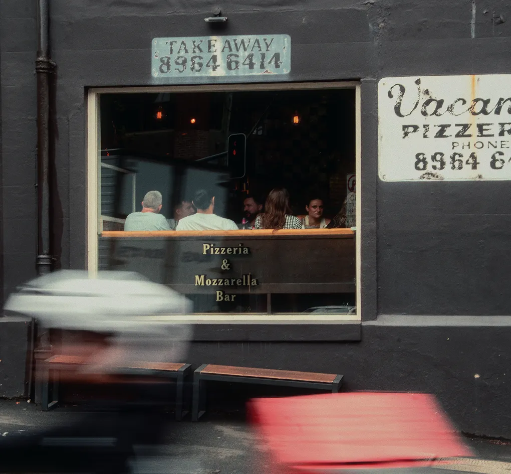 People sitting inside a pizzeria and mozzarella bar seen through a window on a dark wall.