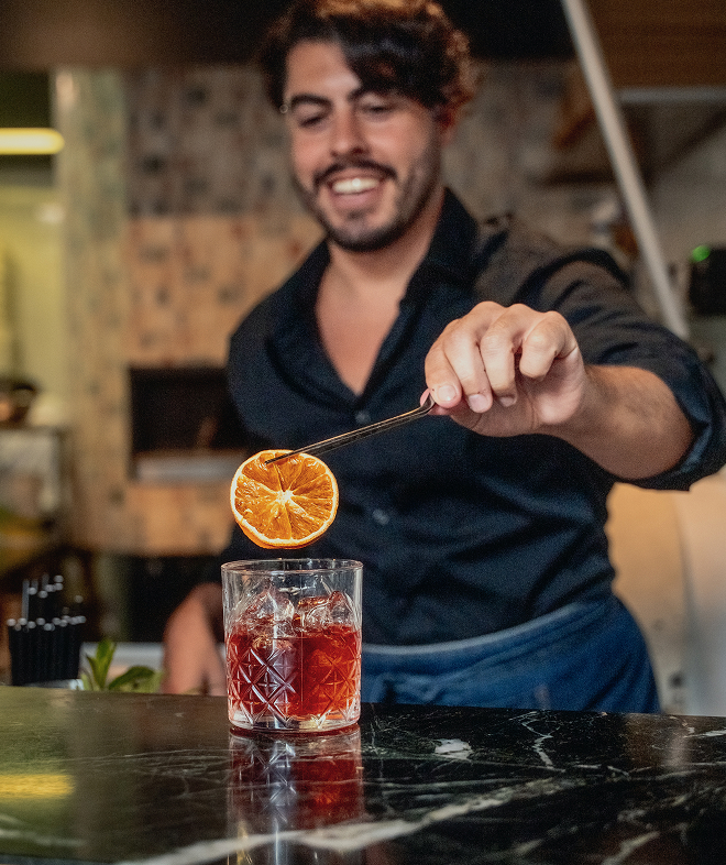 Bartender placing an orange slice garnish on a red cocktail with ice in a patterned glass.