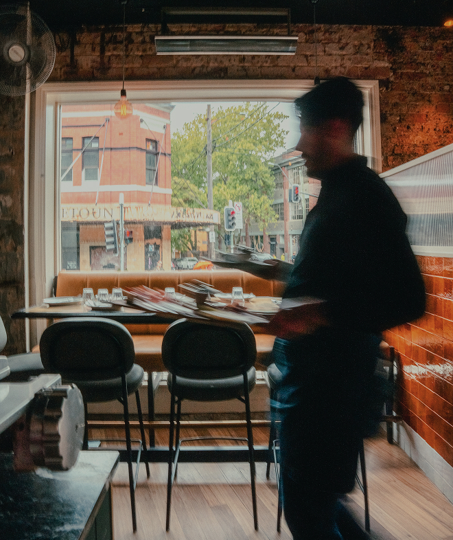 Blurred waiter holding trays in dim restaurant with window showing street and traffic lights outside.