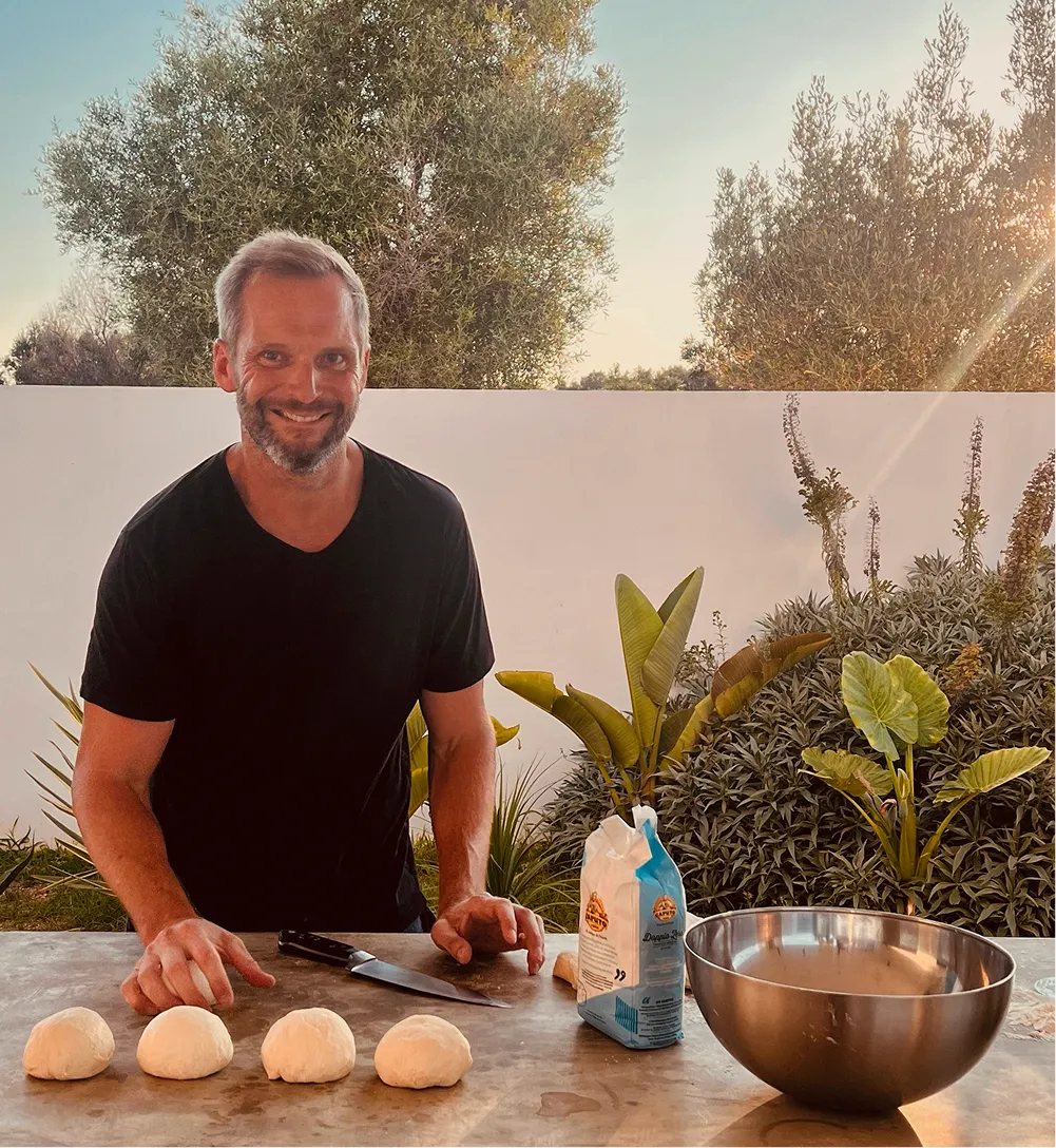 Smiling man in black shirt preparing dough balls on a table with knife, flour, and metal bowl outdoors.