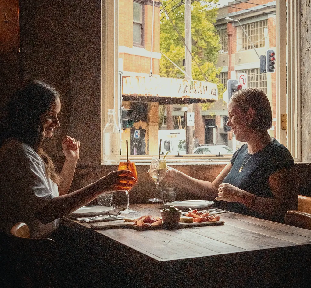Two women sitting at a wooden table by a window, clinking glasses with appetizers on the table.