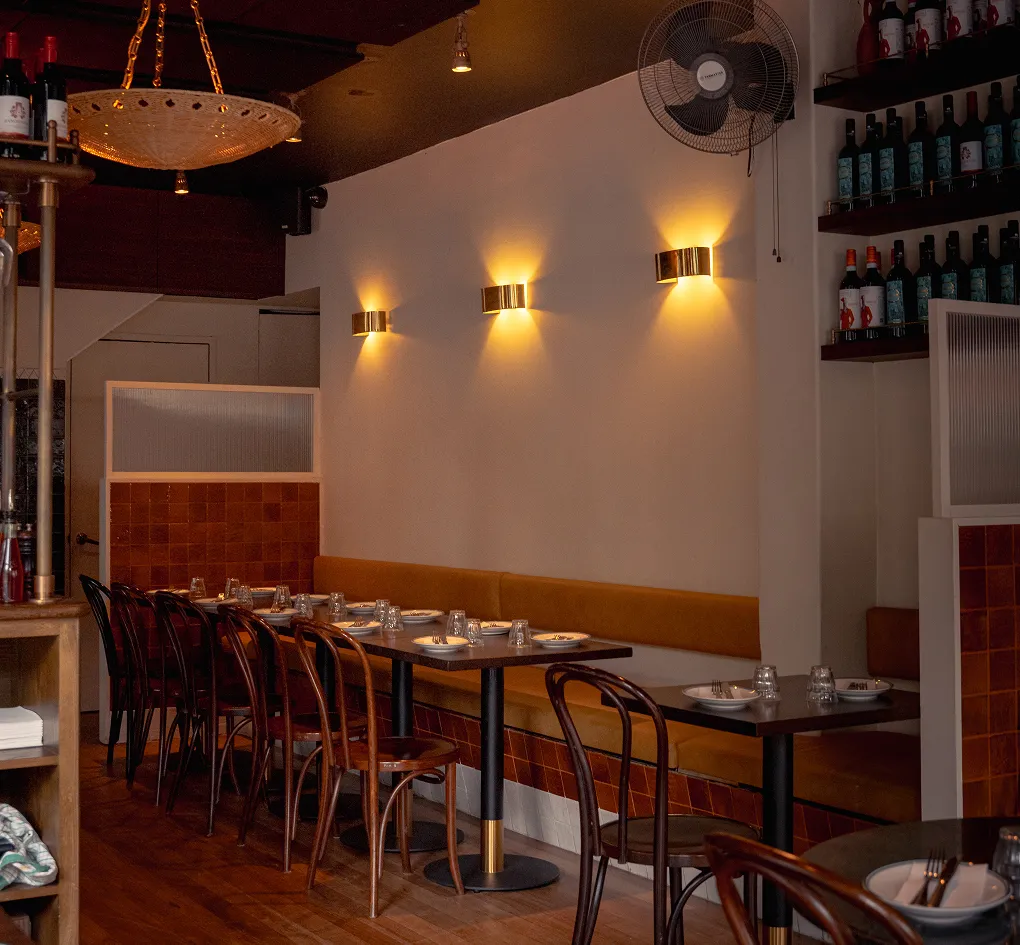 Cozy restaurant interior with wooden chairs, set tables, wall sconces, and bottles on shelves.