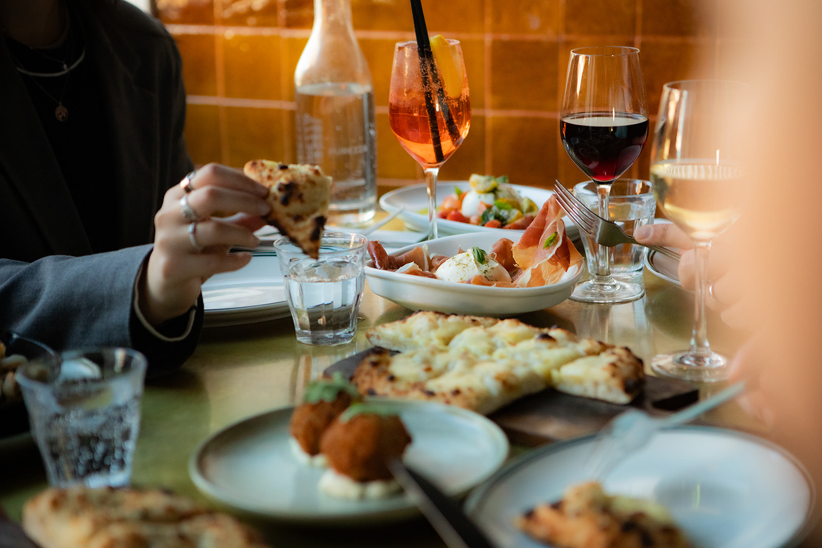 Close-up of a dining table with pizza, appetizers, and glasses of red, white, and orange drinks.