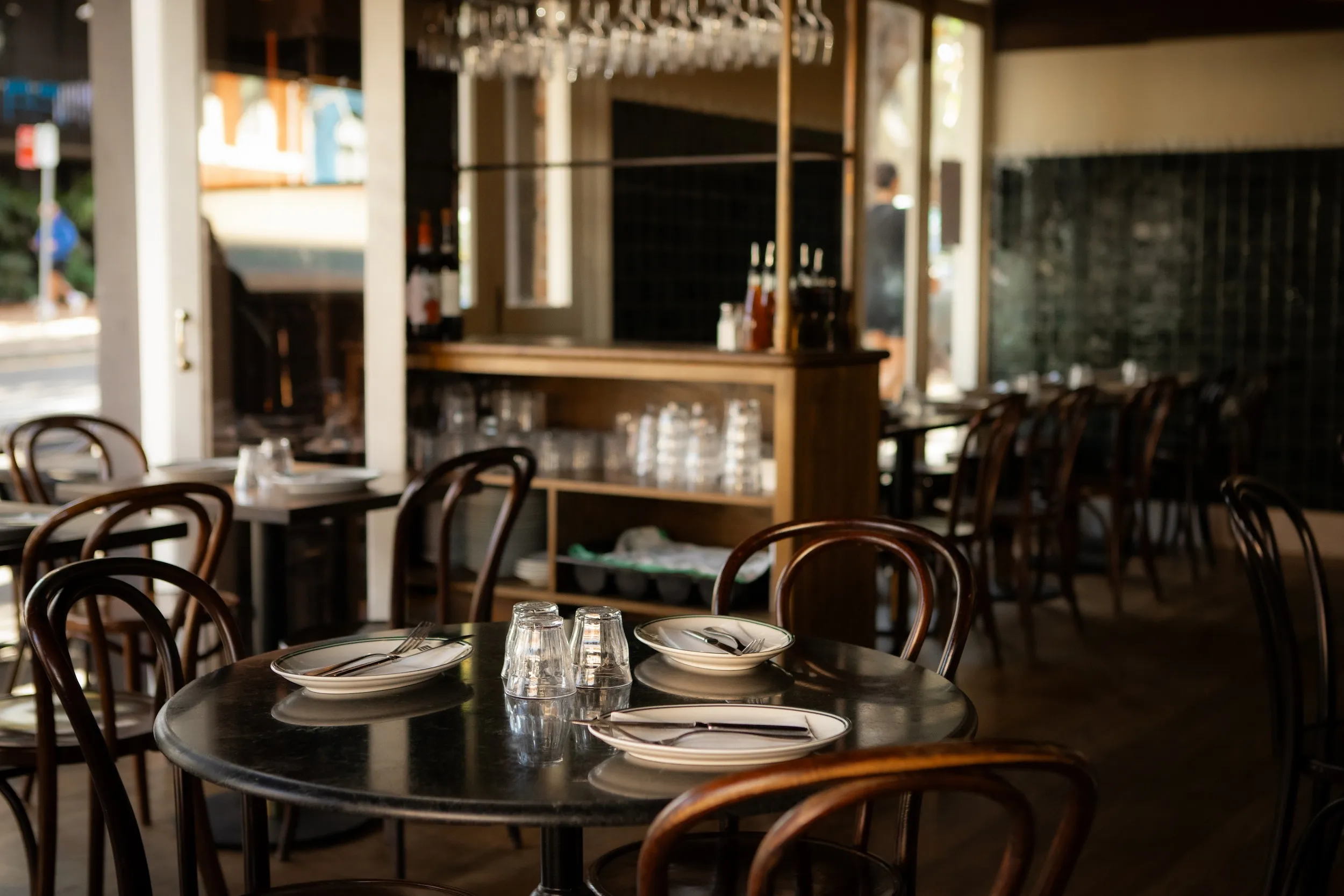 Empty restaurant with wooden chairs, tables set with plates, cutlery, and upside-down glasses.