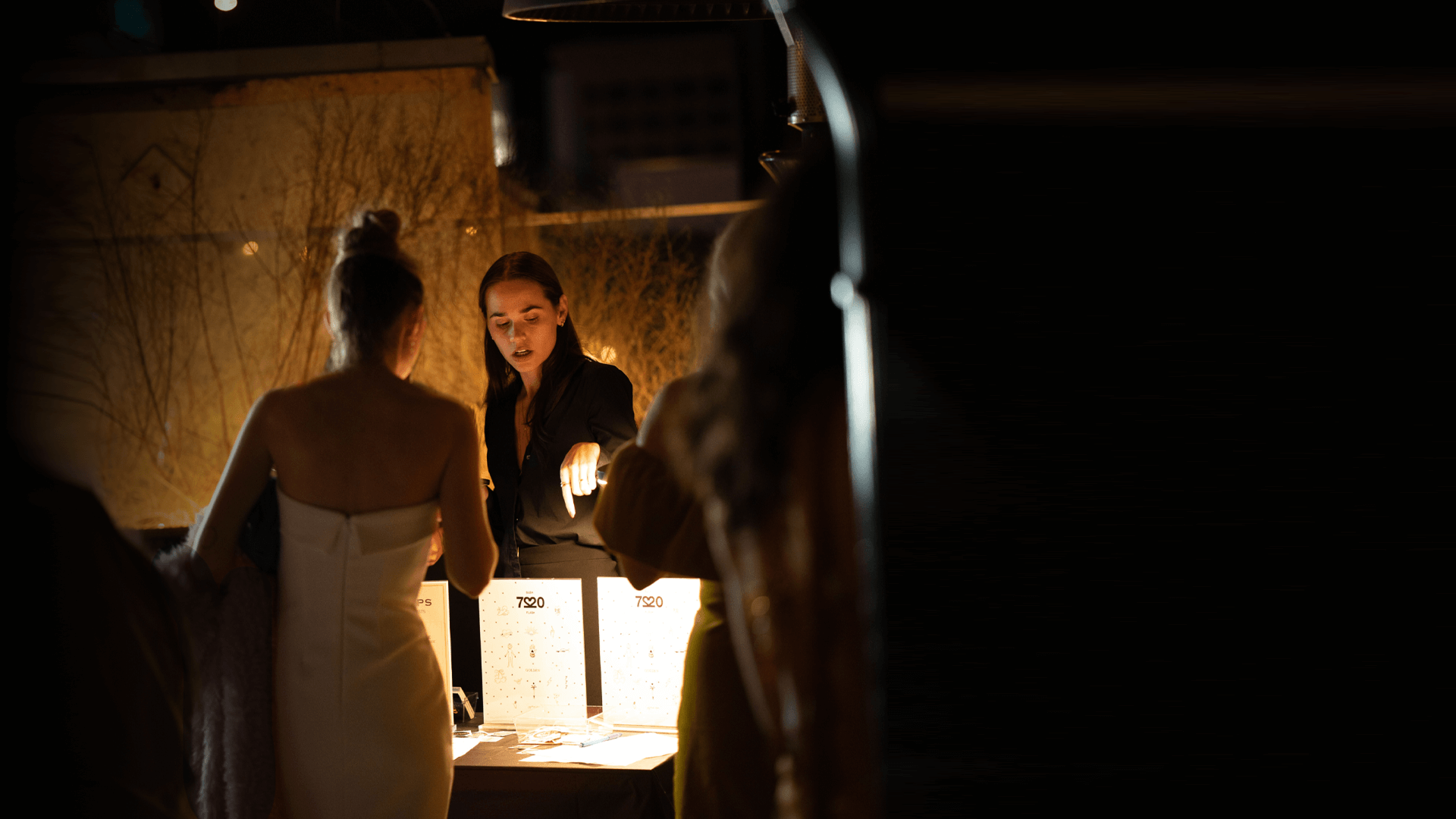Two women at a dimly lit table with signs and one pointing at an item, indoors at night.