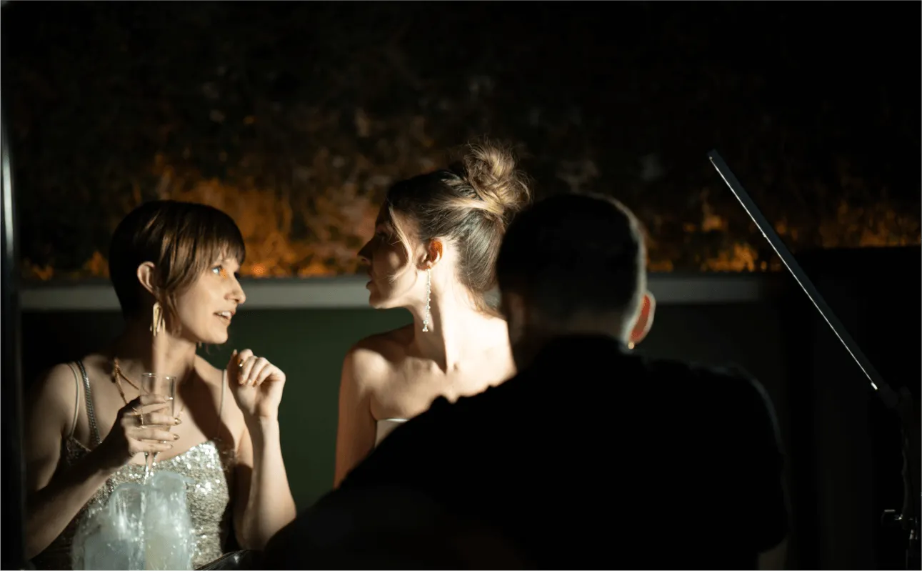 Two women in evening dresses chatting closely, one holding a glass, a man seen from behind nearby.