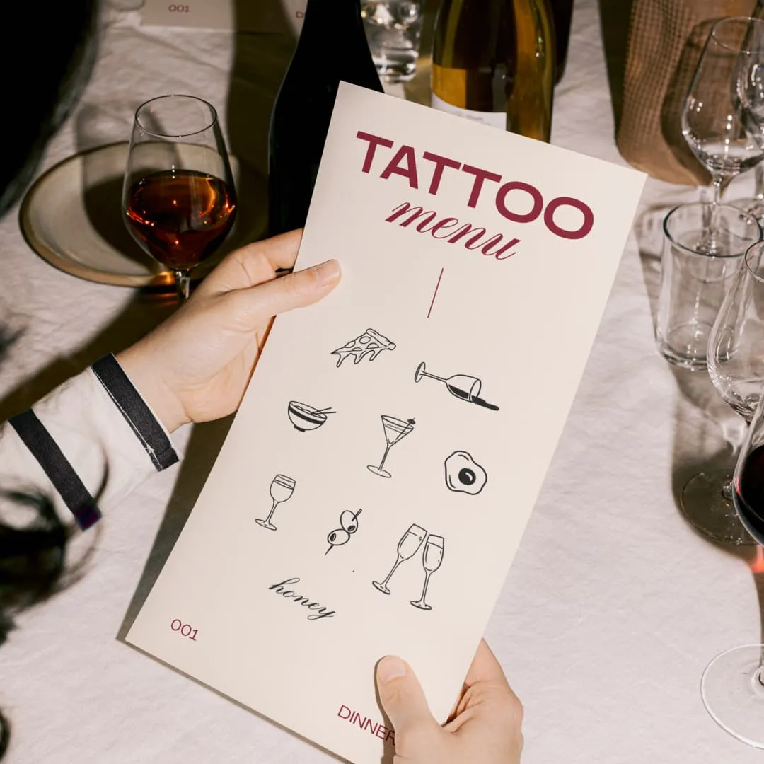 Person holding a menu titled 'Tattoo menu' with food and drink icons on a dinner table.