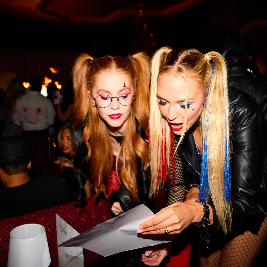 Two women with pigtails in colorful makeup reading a paper in a dimly lit party setting.