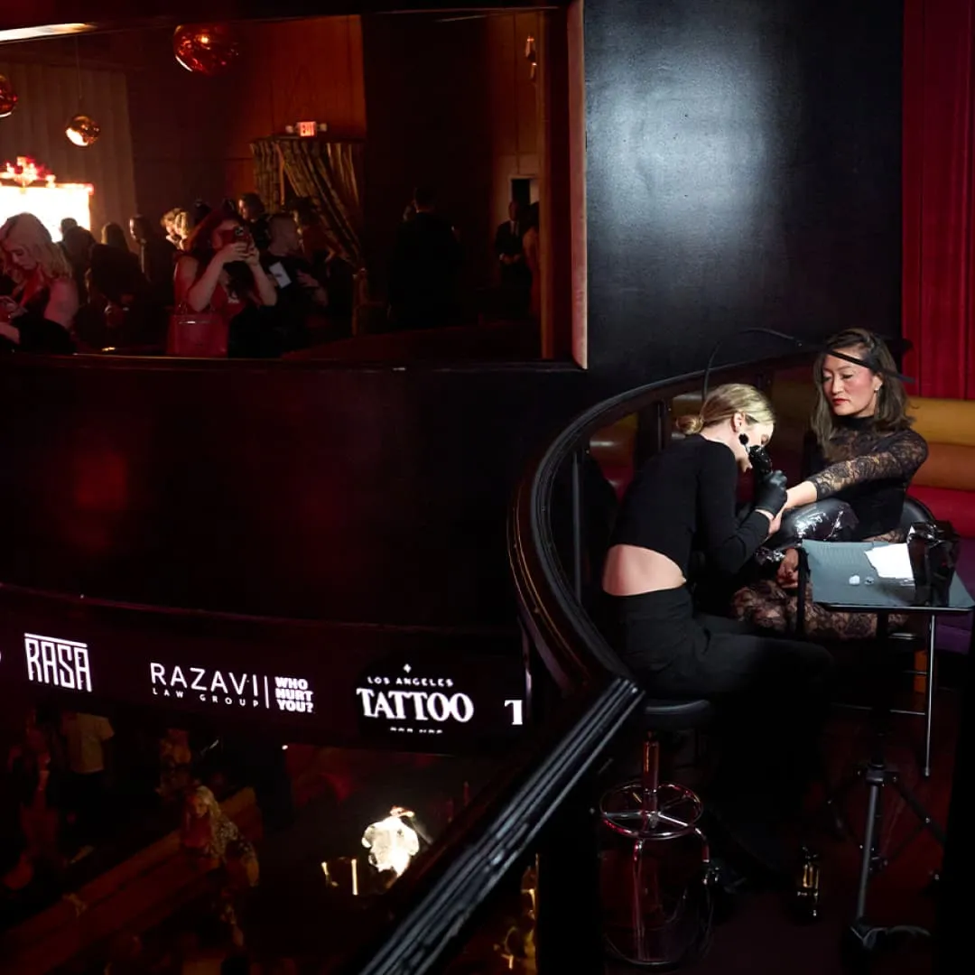 Tattoo artist in black works on a woman's arm at a dimly lit indoor tattoo event.